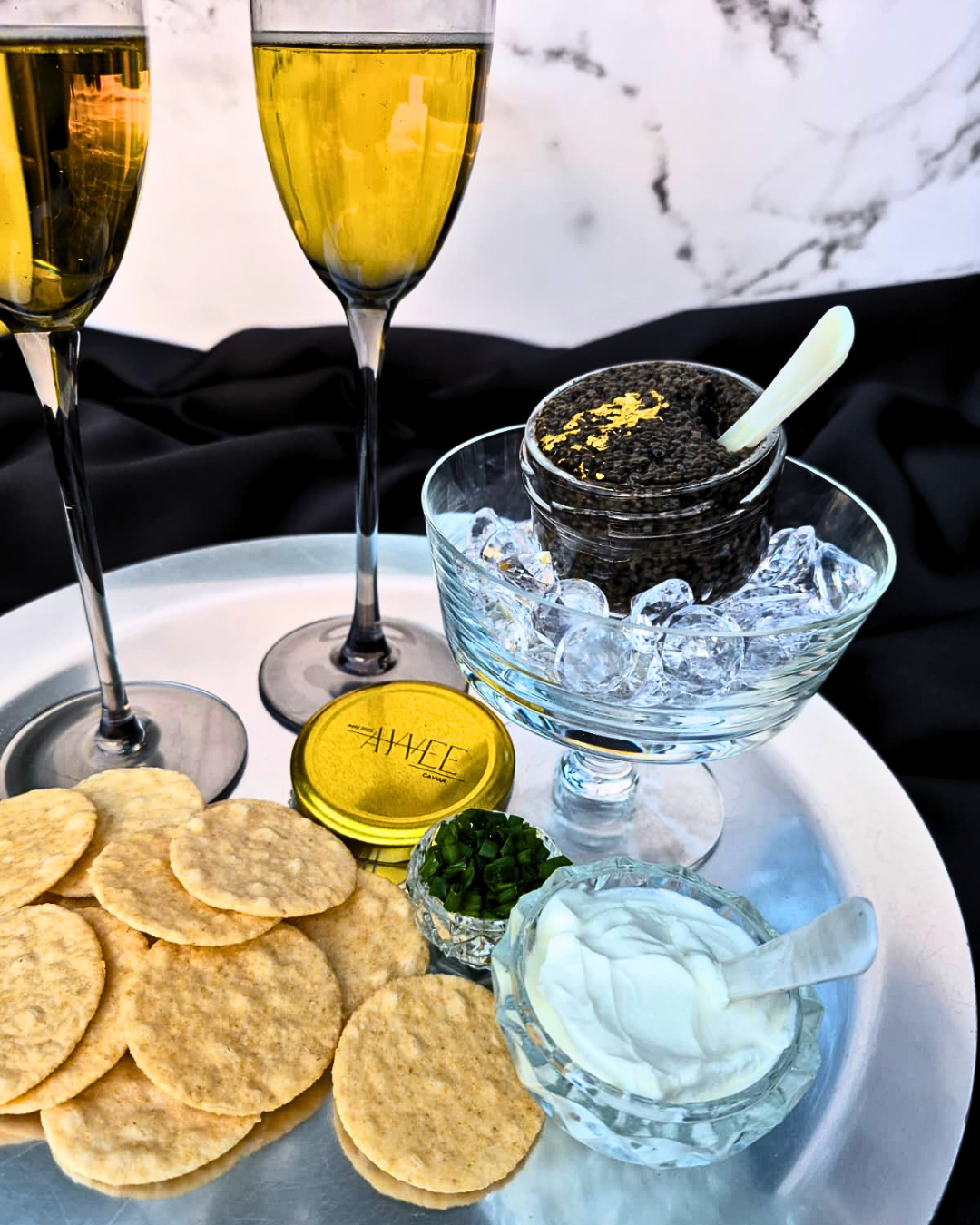 Caviar in a glass bowl with ice, surrounded by crackers and two glasses of champagne on a marble surface.